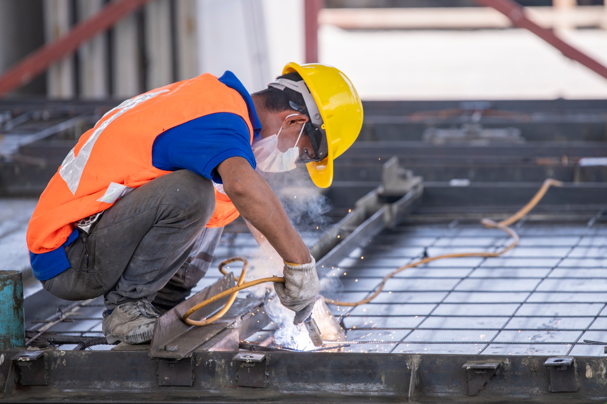 Construction worker engaged in welding work. He is crouching down on a metal framework welding, visi