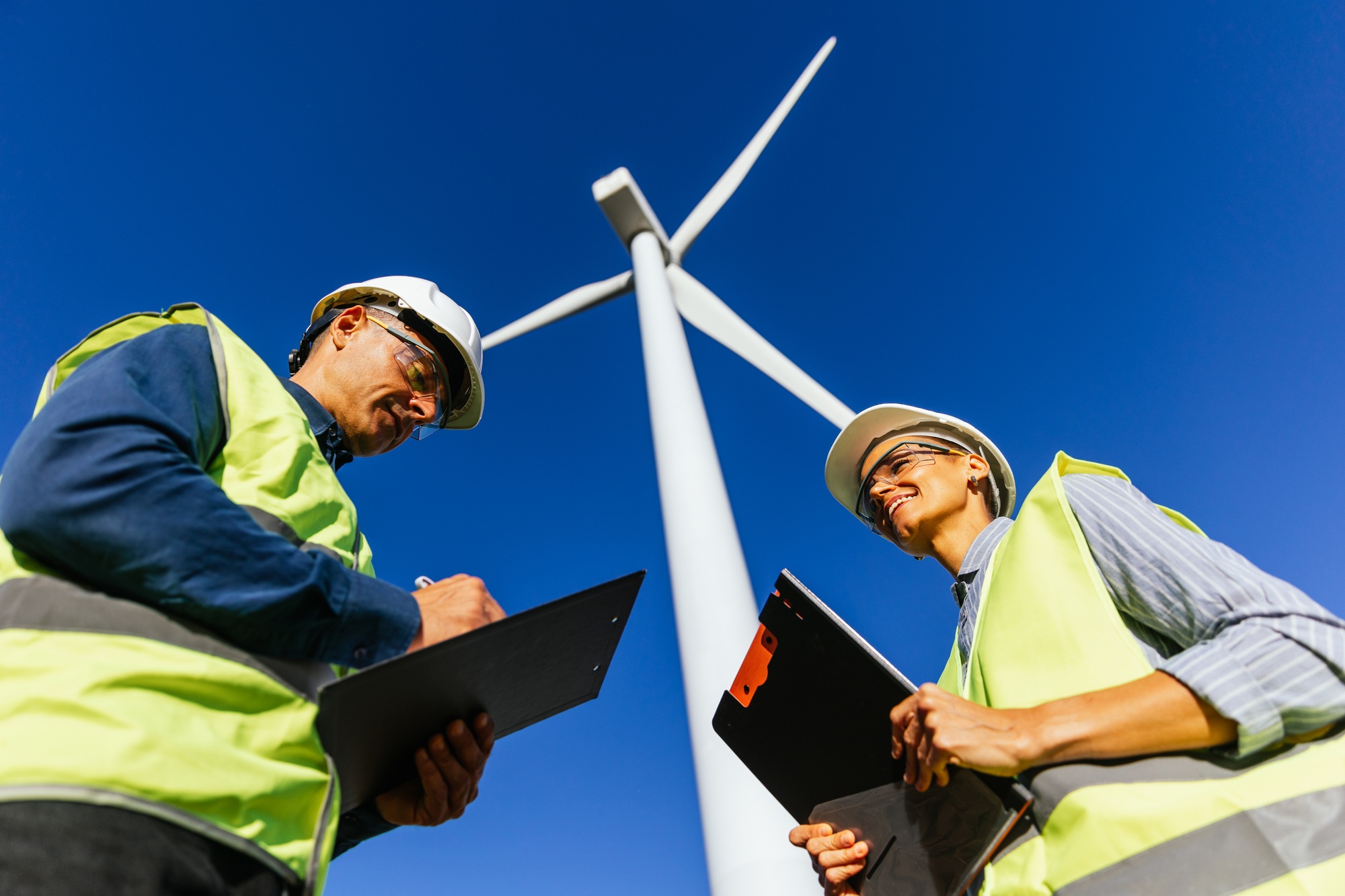 Engineers in helmets and uniforms hold inspection work documents at wind farm.